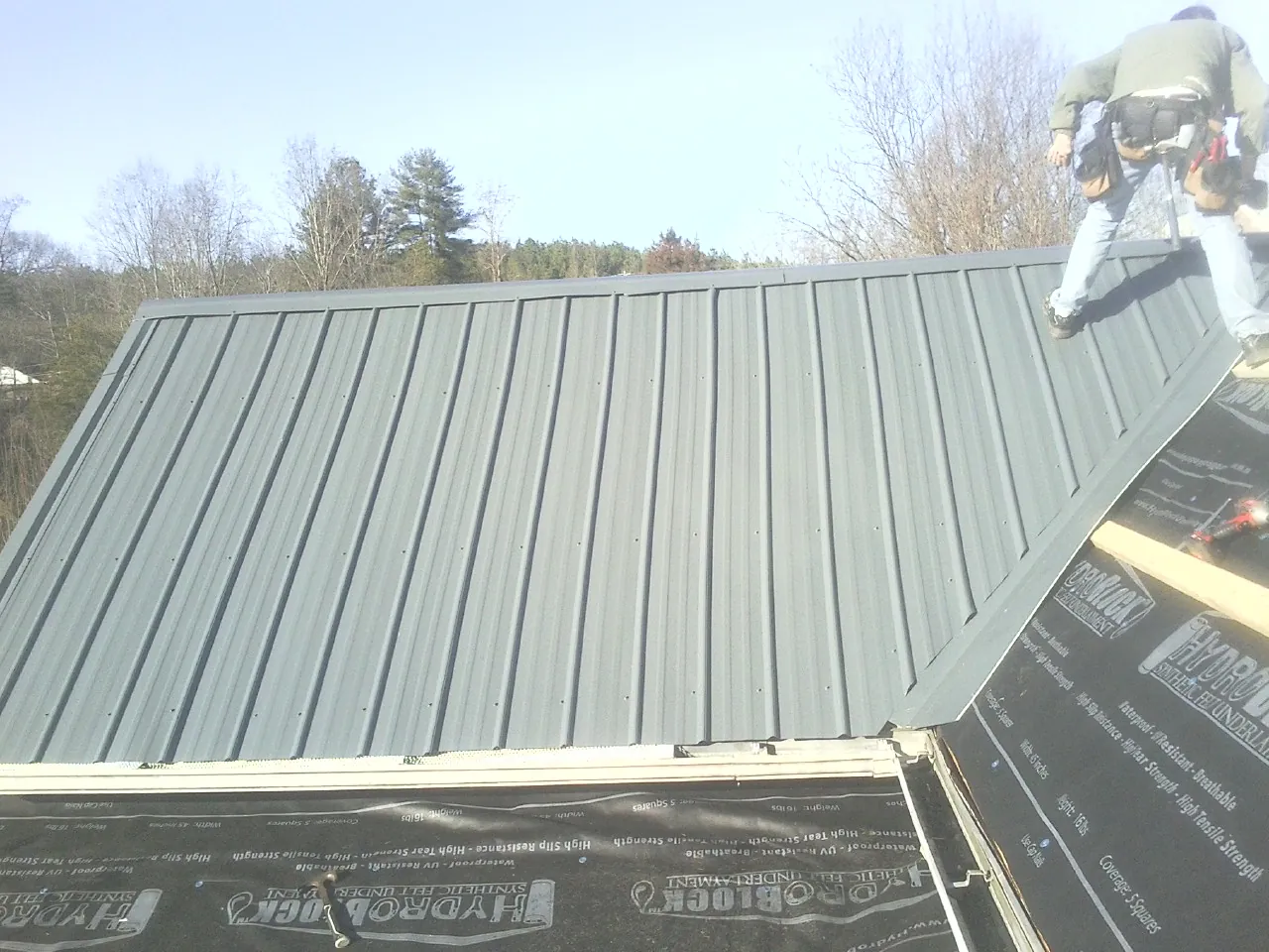 Crew installing dark green standing seam panels over synthetic underlayment in Mossville