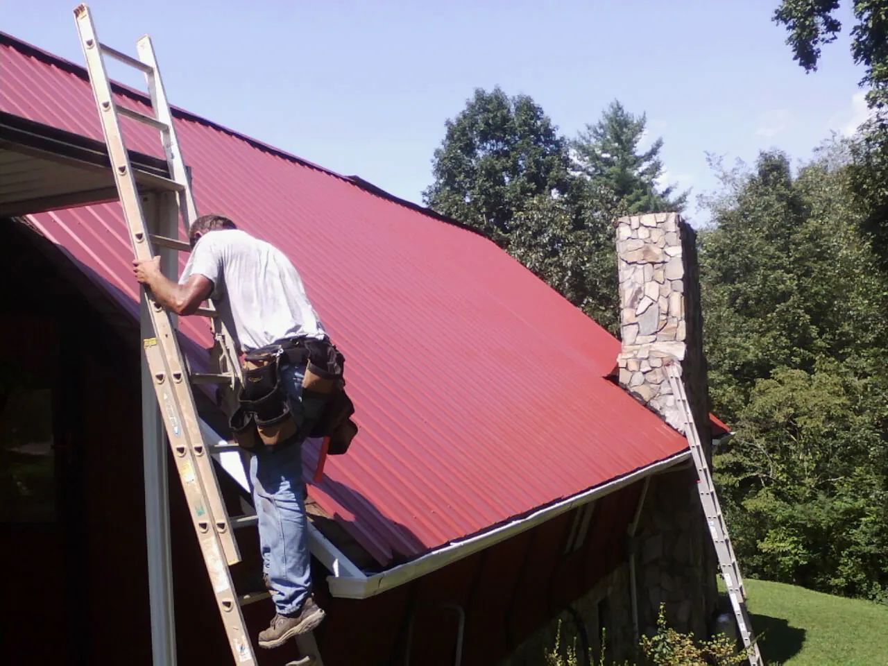 Roofer on a ladder beside a red metal roof and stone chimney — Soffit & Fascia Repair by Roofing Brothers in Mossville